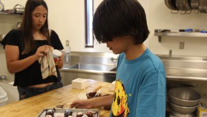 One young person is touching up the bonbons they made while another young person is cleaning a dish in the background.