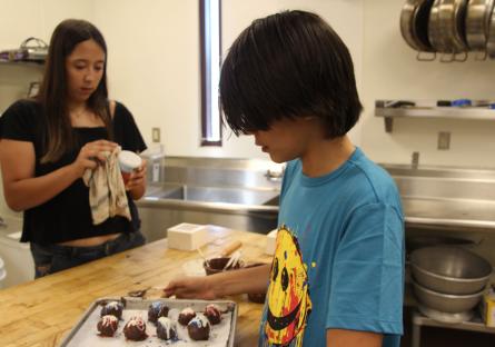 One young person is touching up the bonbons they made while another young person is cleaning a dish in the background.